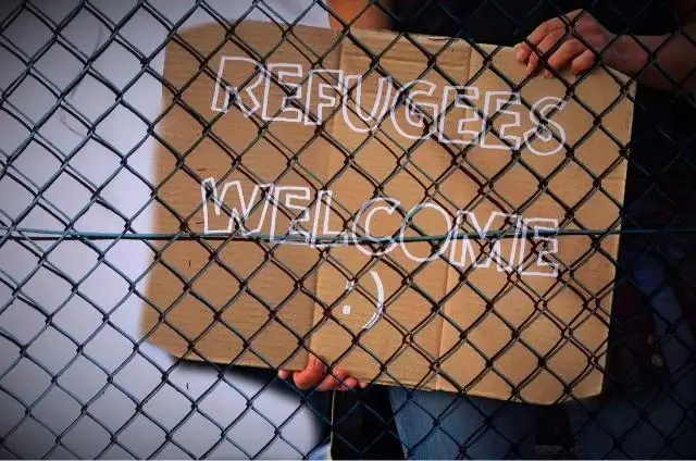 Behind a fence is a sign welcoming refugees